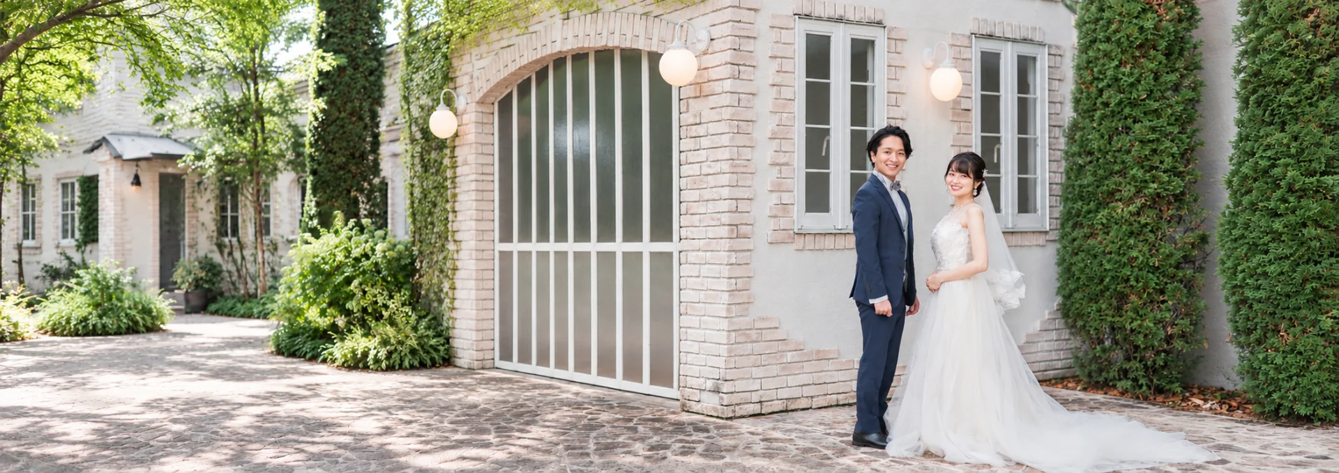 木漏れ日の洋館の前で寄り添い、笑顔で振り向くウェディング姿の男女